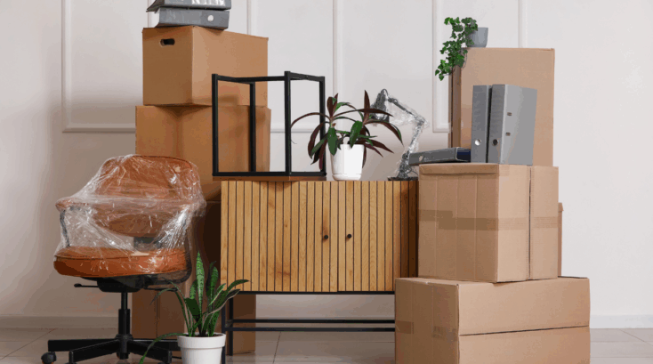 Stacked moving boxes, office chair wrapped in protective film, cabinet, folders and houseplants prepared for storage or moving