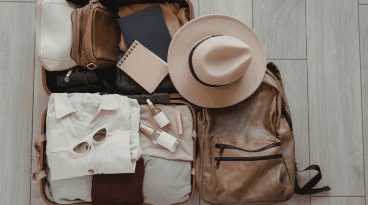 Open suitcase neatly packed with neutral clothing, toiletries, sunglasses and a hat, alongside a leather backpack on a wooden floor.