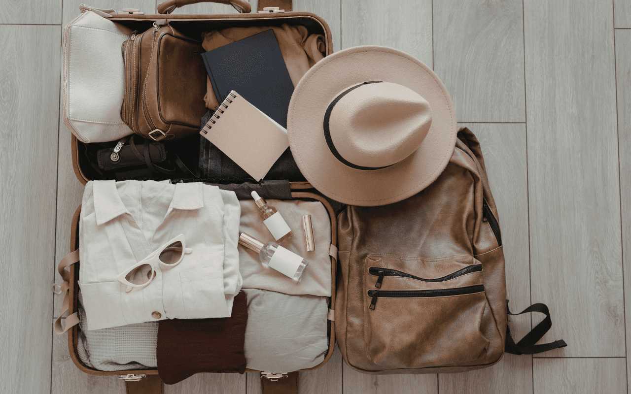 Open suitcase neatly packed with neutral clothing, toiletries, sunglasses and a hat, alongside a leather backpack on a wooden floor.