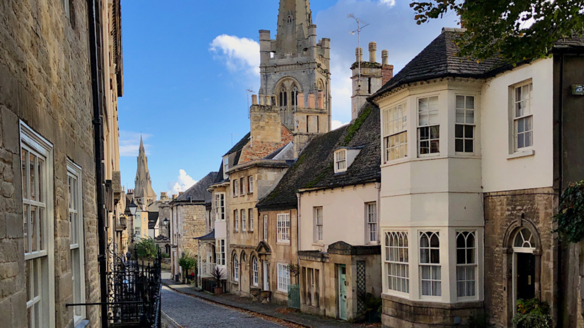 Historic cobbled street in Stamford, Lincolnshire, lined with traditional stone buildings and period homes, with church spires rising above the town under a bright blue sky.