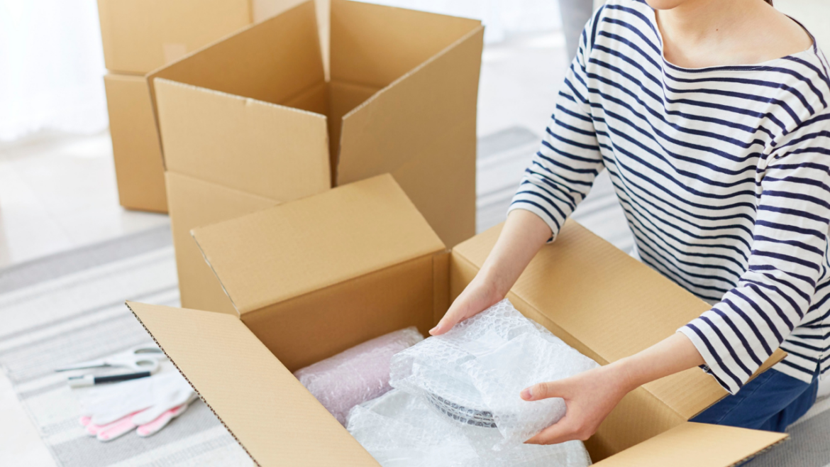 Person carefully packing fragile household items wrapped in bubble wrap into cardboard moving boxes while preparing belongings for storage or a house move.